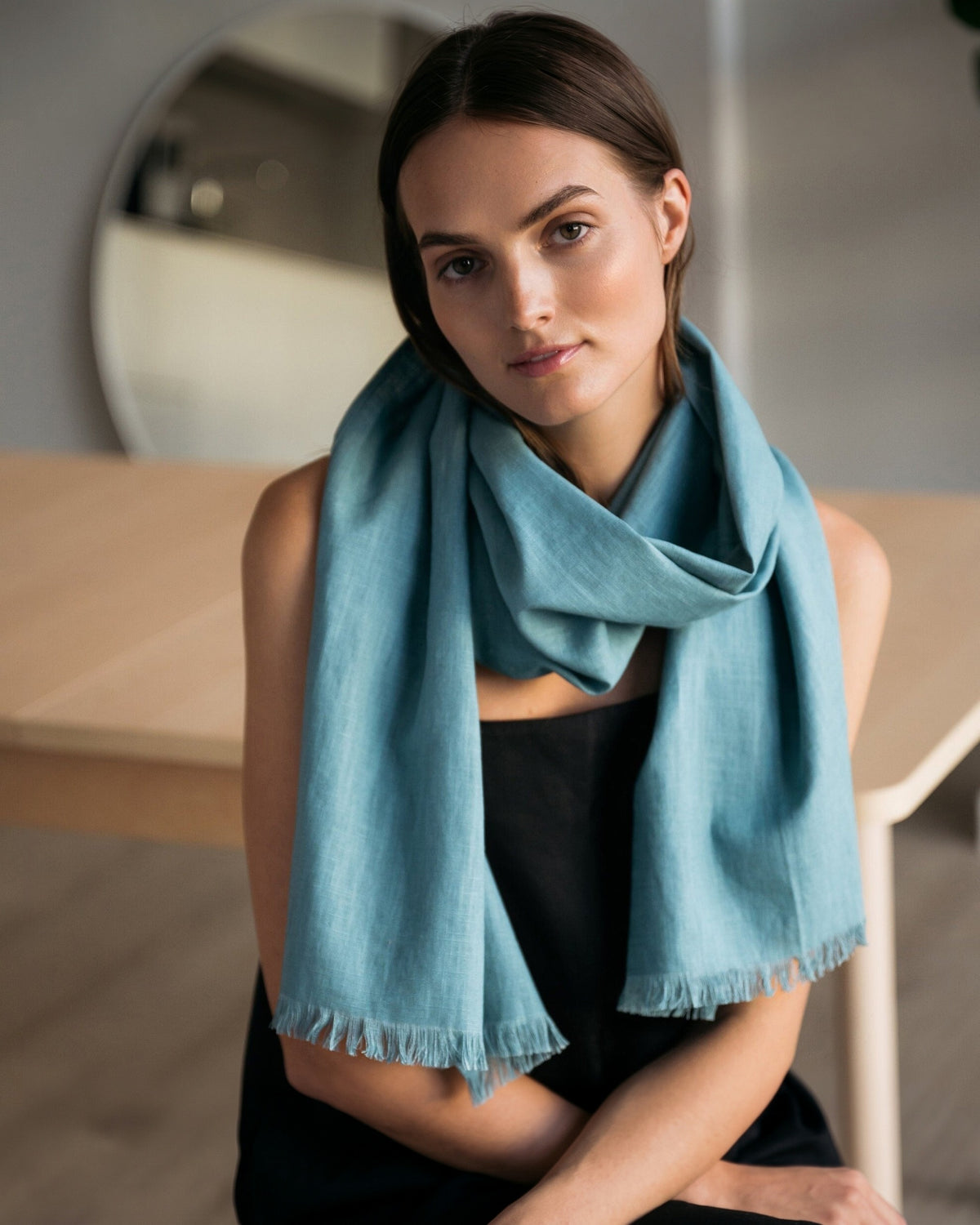 A woman wearing a teal fringed scarf draped over her shoulders, paired with a black top, in a softly lit indoor setting.