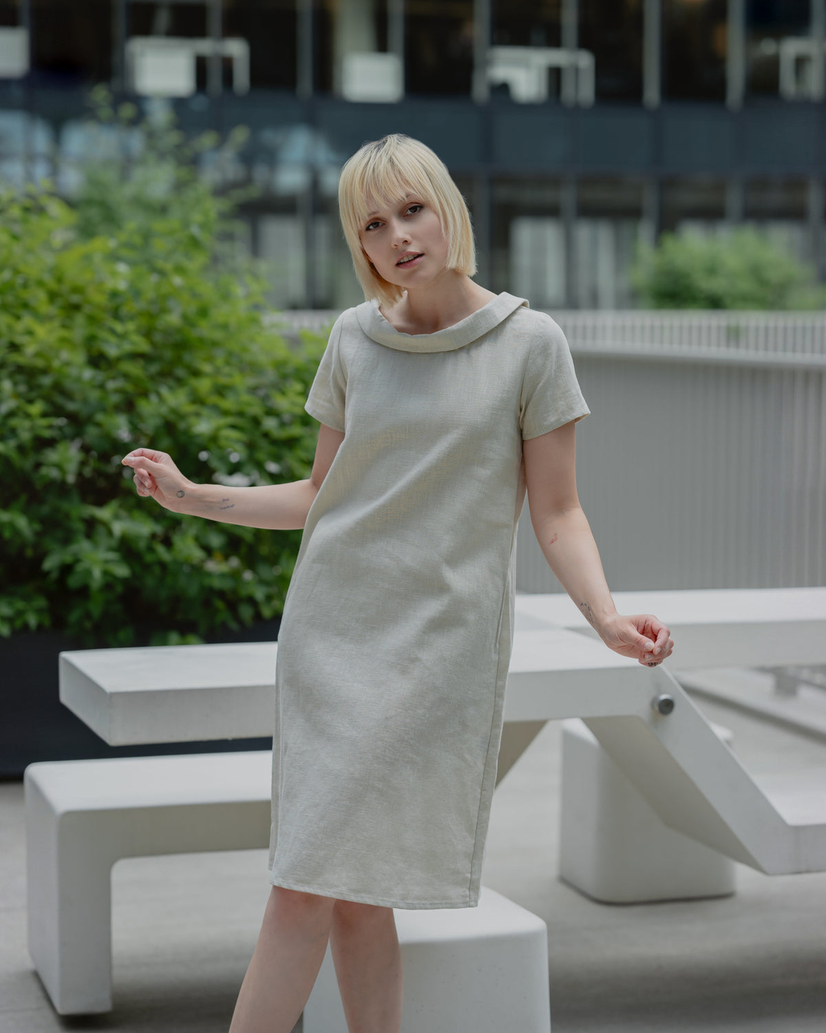 LeMuse STORY linen dress in beige – standing front view, hand resting on a white bench #Color_Beige