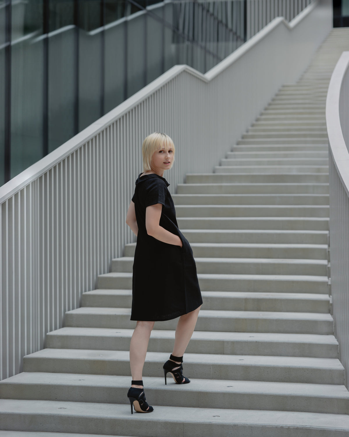 LeMuse STORY linen dress in black – three-quarter back view on stairs, hand in pocket #Color_Black