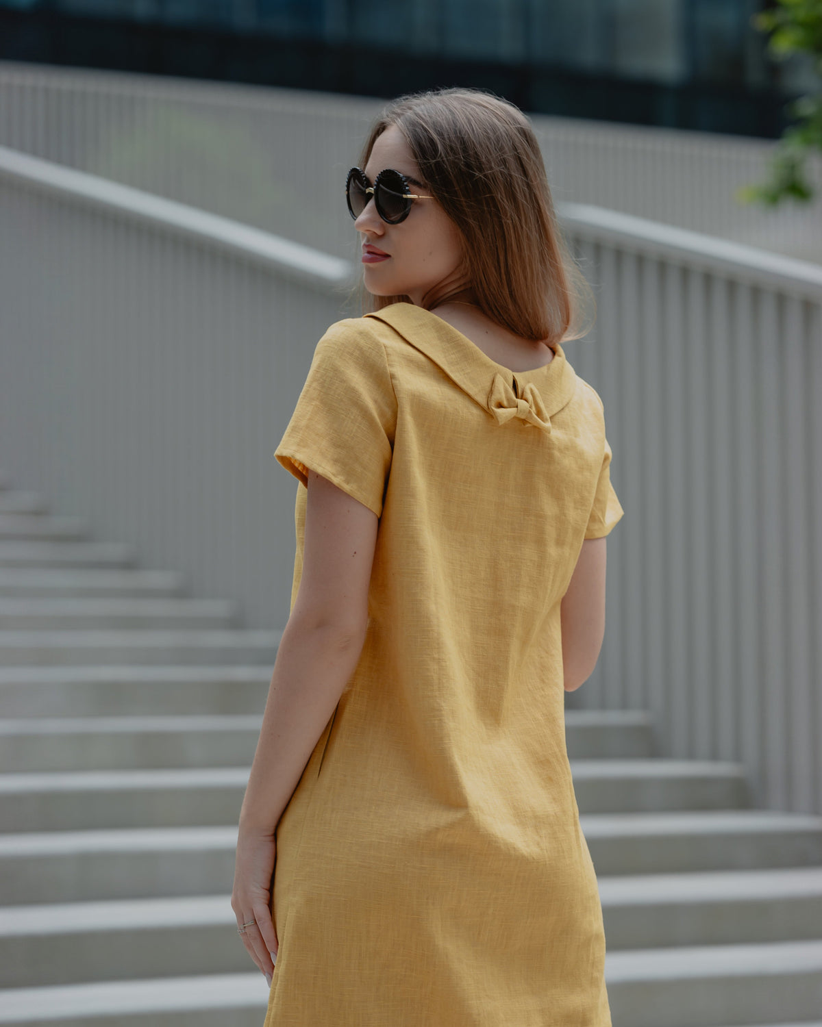 LeMuse STORY linen dress in yellow – standing three-quarter back view on stairs, wearing sunglasses #Color_Yellow
