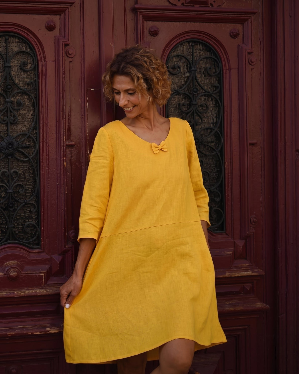 SUN – Linen Blend V-Neck Bow Dress – Yellow dress, three-quarter view holding the hem, at a wooden door #Color_Yellow