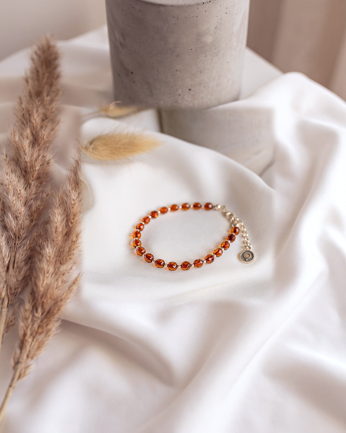 A beaded bracelet with faceted amber-orange beads and a small round charm, displayed on draped white fabric with soft decorative dried grass in the background.